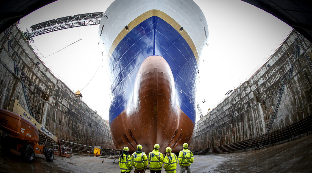 Un navire en cale sèche. Au premier plan, des travailleurs en tenue haute-visibilité regardent vers la